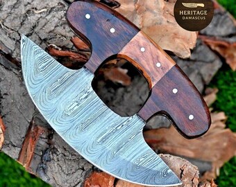 May include: A Damascus steel knife with a curved blade and a dark wood handle. The handle is secured with white rivets. The knife is displayed on a piece of wood. The knife has the text "HERITAGE DAMASCUS" on the handle.