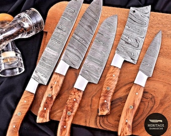 May include: A set of six Damascus steel kitchen knives with wooden handles, displayed on a wooden cutting board. The blades have a distinctive patterned design. A clear glass pepper grinder is visible in the background.