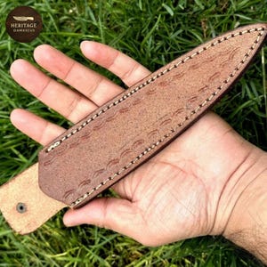 Puede incluir: Una funda de cuchillo de cuero marr&oacute;n con costuras decorativas y patrones de ondas en relieve. La funda se sostiene en una mano sobre un fondo de hierba verde. El logotipo "HERITAGE DAMASCUS" es visible.