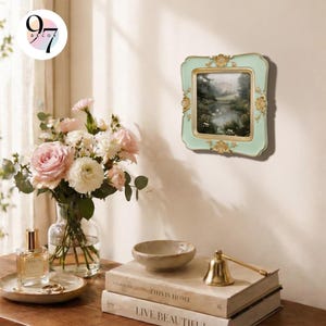 A light green square picture frame with gold ornate details, displaying a landscape painting, hangs on a light wall next to a vase of pink roses.