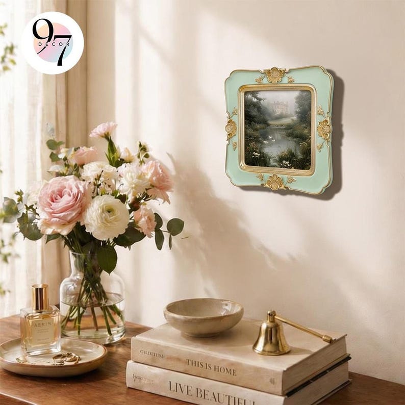 A light green square picture frame with gold ornate details, displaying a landscape painting, hangs on a light wall next to a vase of pink roses.