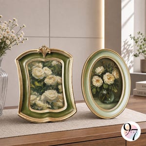 May include: Two decorative picture frames featuring floral artwork. The left frame is rectangular with a scalloped edge, finished in dark green and gold. The right frame is oval, finished in light green and gold. Both contain paintings of white roses.