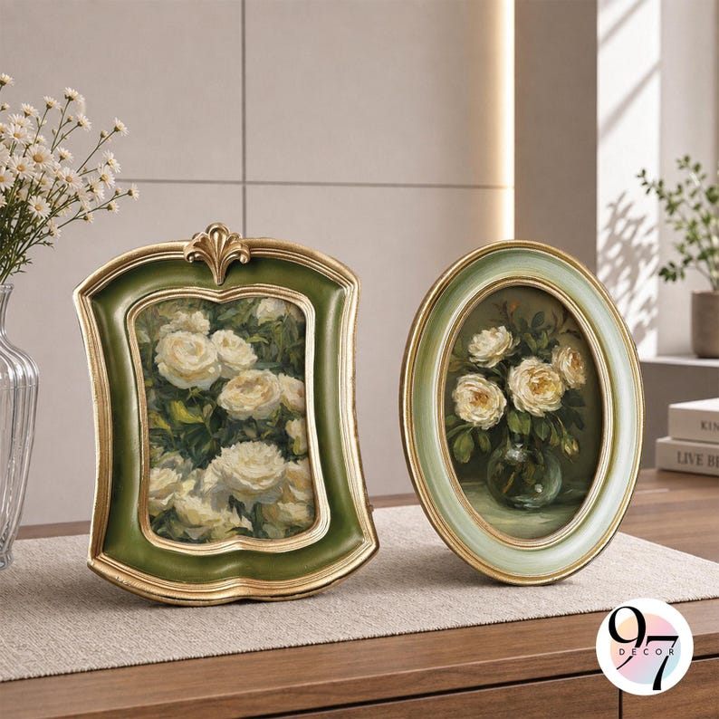 May include: Two decorative picture frames featuring floral artwork. The left frame is rectangular with a scalloped edge, finished in dark green and gold. The right frame is oval, finished in light green and gold. Both contain paintings of white roses.