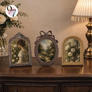 Three ornate picture frames standing on a wooden surface next to a lit lamp and a vase of white flowers.