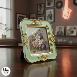 A small, light green square picture frame with gold ornate details, displaying a floral painting, sits on a dark wooden table.