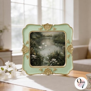 May include: A square picture frame with a light mint green finish and ornate gold-colored floral accents at the center of each side. The frame holds a landscape print depicting a calm pond with white swans, green foliage, and small white flowers.