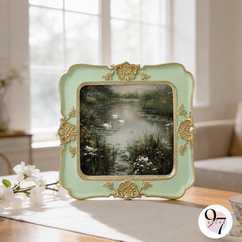 A light green square picture frame with gold ornate corners and edges, displaying a painting of swans on a lake, sits on a white tablecloth.