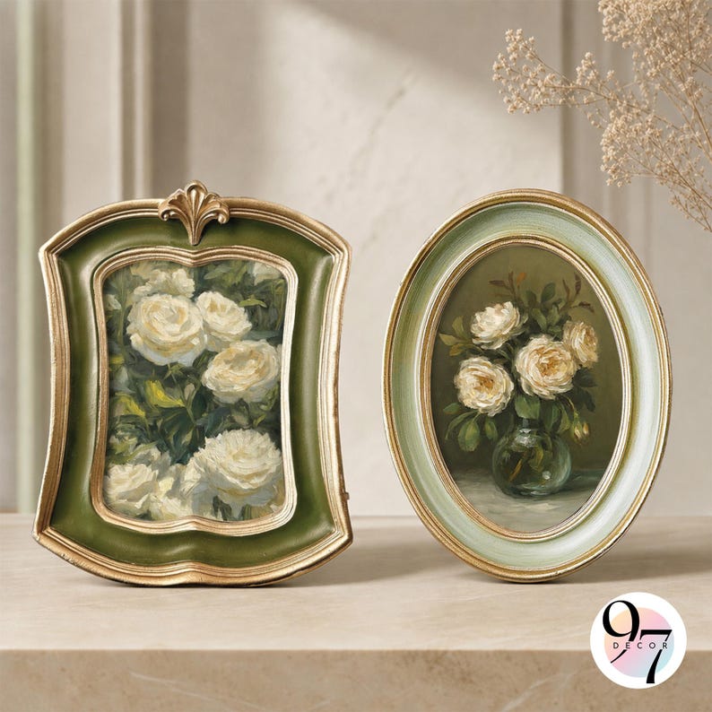 May include: Two decorative frames with floral artwork. The left frame is rectangular with a dark green border and gold trim, displaying white roses. The right frame is oval with a light green border and gold trim, showing white roses in a vase.
