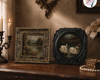 May include: Two ornate picture frames featuring detailed relief carvings. The square frame is finished in a distressed gold tone, while the circular frame is matte black. Both contain landscape paintings, one depicting a river and the other two white swans.