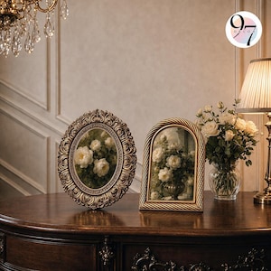 Two ornate picture frames resting upright on a dark wooden surface next to a lit lamp and a vase of white roses.