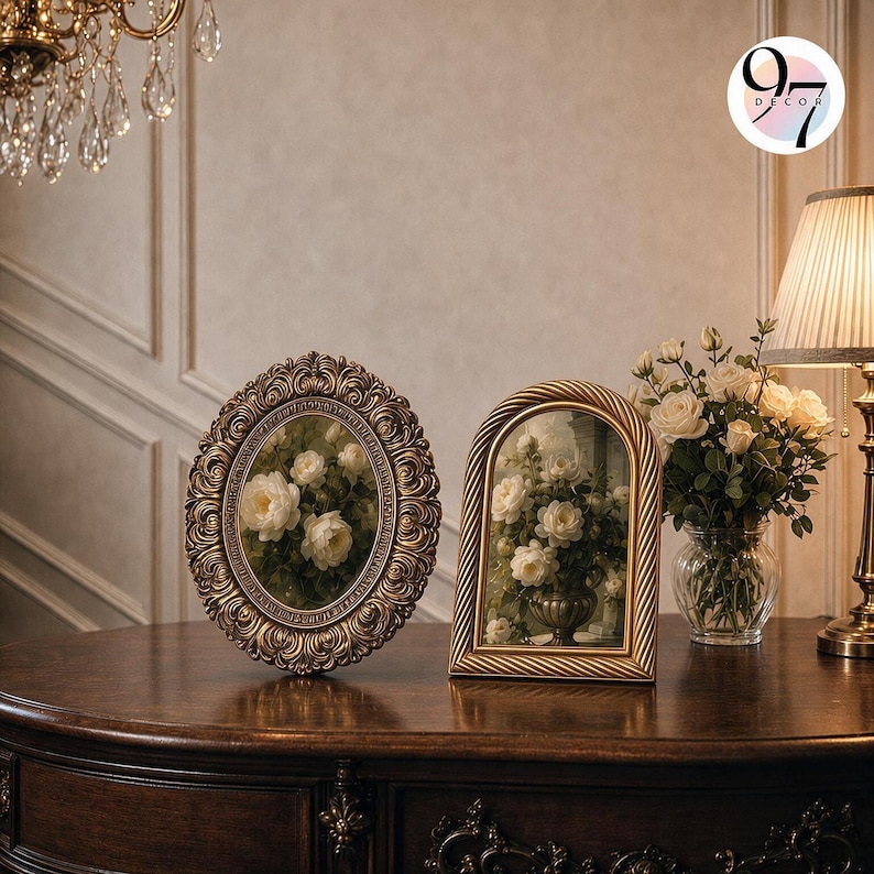 Two ornate picture frames resting upright on a dark wooden surface next to a lit lamp and a vase of white roses.