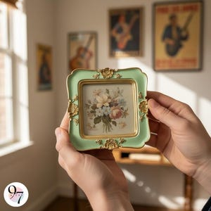 Two hands hold a small, light green square picture frame with gold ornate details, displaying a painting of a floral bouquet.