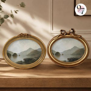 May include: Two oval-shaped gold picture frames containing landscape prints of a lake surrounded by mountains. The left frame features a decorative top detail, while the right frame is adorned with a prominent gold bow at the top centre.
