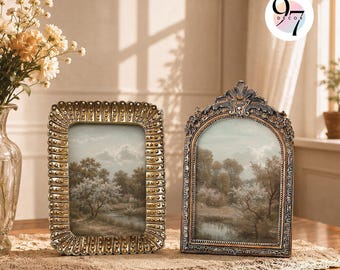 May include: Two ornate picture frames with metallic gold finishes. The left frame is rectangular with a beaded border, while the right frame features an arched top with intricate scrollwork. Both contain landscape prints of trees and a river.