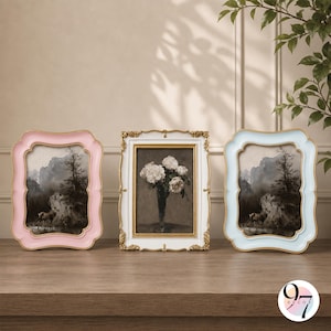 May include: Three rectangular picture frames standing on a wooden surface. The left frame is light pink, the right is light blue, both with smooth edges. The center frame is white with gold ornate detailing, containing a print of white flowers.