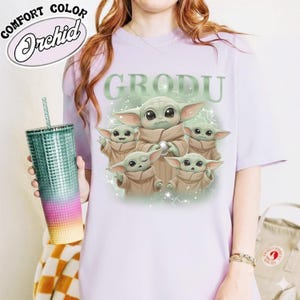 May include: A light purple t-shirt featuring a graphic of Grogu, with the word "GRODU" above the image. The design includes five Grogu characters in various poses. A teal tumbler with a straw is held in the left hand.
