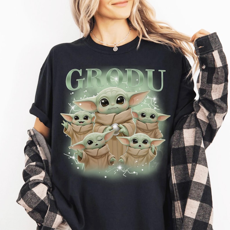 May include: Black t-shirt featuring a graphic of Grogu, also known as Baby Yoda, surrounded by four smaller versions of himself. The word "GRODU" is printed above the image in a light green color. The design has a cartoon style.