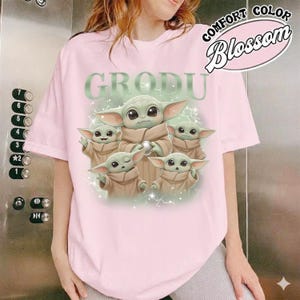 May include: A light pink t-shirt featuring a graphic of Grogu, with the word "GRODU" above the image. The graphic includes multiple Grogu characters in tan robes. The shirt also has the words "COMFORT COLOR Blossom" in a pink oval.