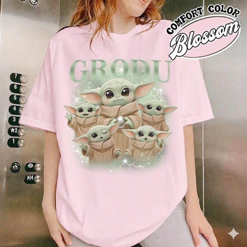 May include: A light pink t-shirt featuring a graphic of Grogu, with the word "GRODU" above the image. The graphic includes multiple Grogu characters in tan robes. The shirt also has the words "COMFORT COLOR Blossom" in a pink oval.