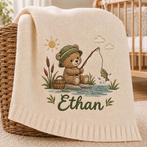 Könnte beinhalten: Eine cremefarbene Strickdecke für Babys mit einer gestickten Szene eines braunen Teddybären, der einen grünen Hut trägt und mit einer Angel fischt. Das Design umfasst eine Sonne, Wolken, Schilf, einen Korb und einen Fisch. Der Name Ethan ist in Grün gestickt.