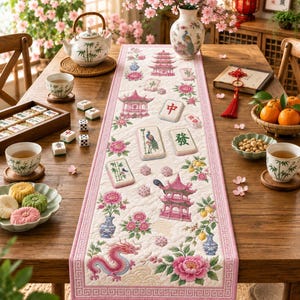 May include: A quilted table runner featuring a pink border and a repeating pattern of pink pagodas, dragons, peonies, and mahjong tiles. The runner is laid out on a wooden table alongside a tea set, a bowl of nuts, oranges, and a game set.