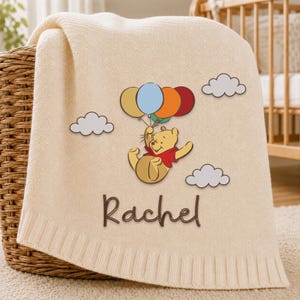 May include: A cream-colored knit baby blanket featuring a graphic of Winnie the Pooh holding a cluster of five colorful balloons, surrounded by three white clouds. The name Rachel is embroidered in brown script lettering below the character.