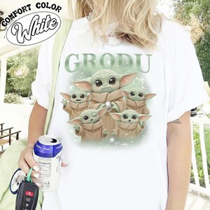 May include: White t-shirt featuring a graphic of Grogu, also known as Baby Yoda, from The Mandalorian. The design includes Grogu in various poses with the word "GRODU" above. The shirt has the words "COMFORT COLOR" and "White" in a circular design.