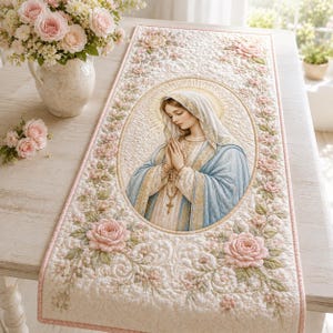 May include: A rectangular quilted table runner featuring a central oval portrait of the Virgin Mary with hands clasped in prayer, surrounded by a border of embroidered pink roses, green leaves, and a soft cream-colored quilted fabric background.