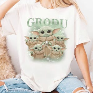 May include: White t-shirt featuring a graphic of Grogu, with the word "GRODU" above the image. The graphic shows Grogu surrounded by four smaller versions of himself, all in tan robes, with a light green background.