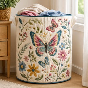 May include: A cylindrical quilted laundry hamper in cream fabric featuring embroidered colorful butterflies and floral patterns in shades of pink, blue, and yellow. The hamper has a teal trim, two side handles, and contains folded fabric items.