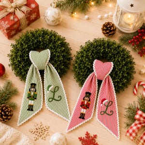 May include: Two circular green boxwood wreaths featuring decorative fabric ribbons. One ribbon is green-striped with a nutcracker and the letter L, the other is red-striped with a nutcracker and the letter L. Both ribbons have scalloped white edges.