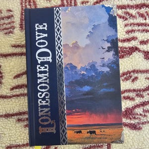 May include: A hardcover book titled "Lonesome Dove" with a navy blue spine and a landscape painting on the cover. The cover art depicts a sunset over a field with cattle. The title is in silver lettering.
