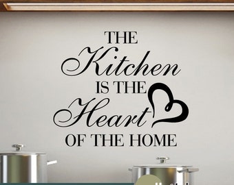 May include: Black and white vinyl wall decal with the text "The Kitchen is the Heart of the Home" and a heart shape. The decal is displayed on a white wall above a kitchen counter.