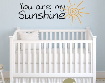 Peut inclure: Un lit bébé blanc avec un matelas blanc et un oreiller blanc. Le mur derrière le lit bébé est bleu clair et comporte un autocollant mural noir et orange qui dit "You are my Sunshine" avec un graphique de soleil.
