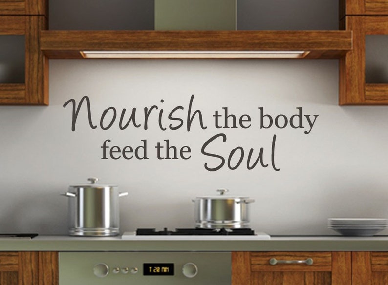 May include: A kitchen with a white wall and a wooden cabinet above the stove. The wall has a black vinyl decal with the text "Nourish the body feed the Soul".