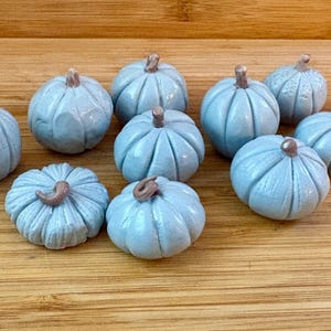 May include: A collection of ten small, light blue ceramic pumpkins with brown stems. The pumpkins are arranged on a wooden surface, showcasing their ribbed texture and autumnal theme. Each pumpkin is approximately 2 inches in diameter.