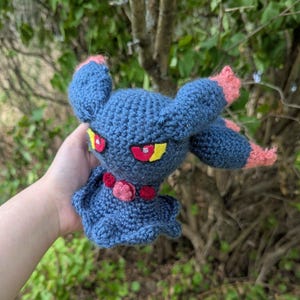 Puede incluir: Muñeca de crochet hecha a mano con forma de criatura azul oscuro con detalles rosas en las orejas y los brazos. La muñeca tiene ojos rojos con detalles amarillos y un collar floral rojo y rosa.