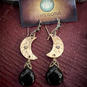 May include: A pair of gold-toned crescent moon earrings featuring a hammered texture and a small stamped heart. Each earring is accented with a wire-wrapped black teardrop-shaped stone and hangs from a hook. The display card reads GYPSYLOTUS.