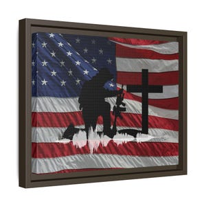 May include: A framed canvas print featuring an American flag with a silhouette of a kneeling soldier and a cross. The flag's red and white stripes and blue field with stars are prominently displayed. The soldier is holding a rifle.