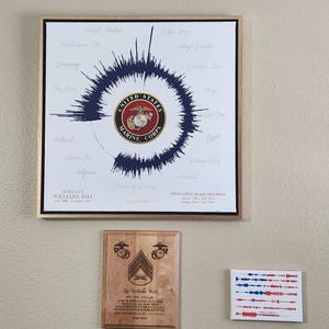 May include: A framed print featuring a navy blue sound wave design around the United States Marine Corps emblem. Below, a wooden plaque with a Marine Corps insignia and text. A small canvas with a red, white, and blue sound wave design is also visible.
