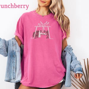 May include: A bright pink t-shirt with a white graphic of a group of people around a table. The word "Crunchberry" is printed in pink at the top left. The shirt is a loose fit and has short sleeves.