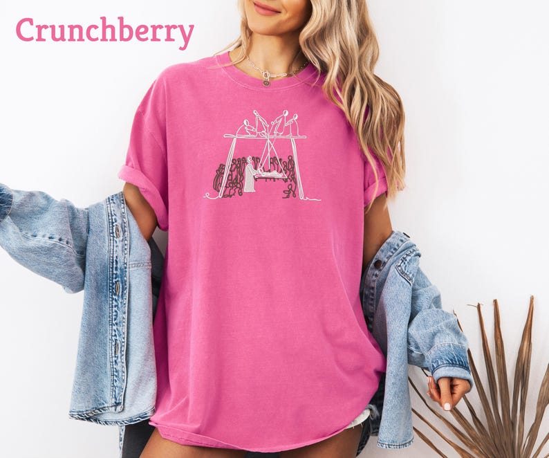 May include: A bright pink t-shirt with a white graphic of a group of people around a table. The word "Crunchberry" is printed in pink at the top left. The shirt is a loose fit and has short sleeves.