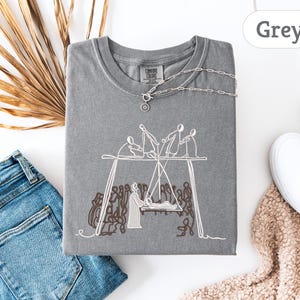 May include: A grey t-shirt with a crew neck and short sleeves. The shirt features a white embroidered design of figures on a swing set. A silver necklace with a pendant is on the shirt. Blue jeans and white sneakers are also in the image.