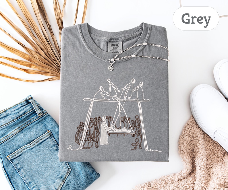 May include: A grey t-shirt with a crew neck and short sleeves. The shirt features a white embroidered design of figures on a swing set. A silver necklace with a pendant is on the shirt. Blue jeans and white sneakers are also in the image.