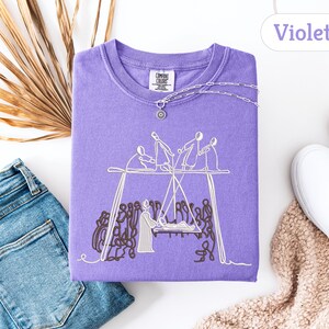 May include: A violet Comfort Colors sweatshirt with an embroidered design of figures on a swing set. The sweatshirt is paired with blue jeans, white sneakers, and a necklace. The word "Violet" is displayed in a white oval.