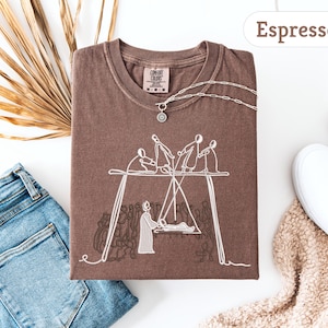 May include: Espresso-colored t-shirt with a white graphic of figures on a swing set. The shirt is folded, with a silver necklace and a Comfort Colors tag visible. A pair of blue jeans and white sneakers are in the background.