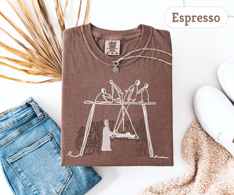 May include: Espresso-colored t-shirt with a white graphic of figures on a swing set. The shirt is folded, with a silver necklace and a Comfort Colors tag visible. A pair of blue jeans and white sneakers are in the background.