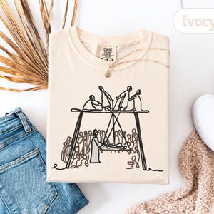 May include: Ivory-colored t-shirt with a black line drawing of a group of people on a stage. The shirt is folded and surrounded by a pair of blue jeans, white shoes, and a dried palm leaf.