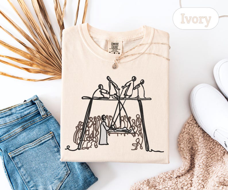 May include: Ivory-colored t-shirt with a black line drawing of a group of people on a stage. The shirt is folded and surrounded by a pair of blue jeans, white shoes, and a dried palm leaf.