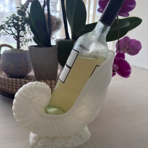 May include: A white, seashell-shaped wine bottle holder cradles a clear wine bottle filled with yellow liquid. The holder has a textured surface and sits on a white surface. The bottle has a black cap and a white label.